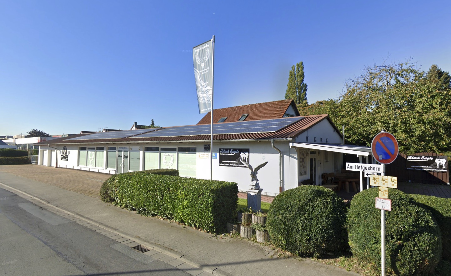 Straßenansicht der Trainingshalle: weißes Gebäude mit Black-Eagle-Beschilderung, Vereinsfahne und Adler-Statue.